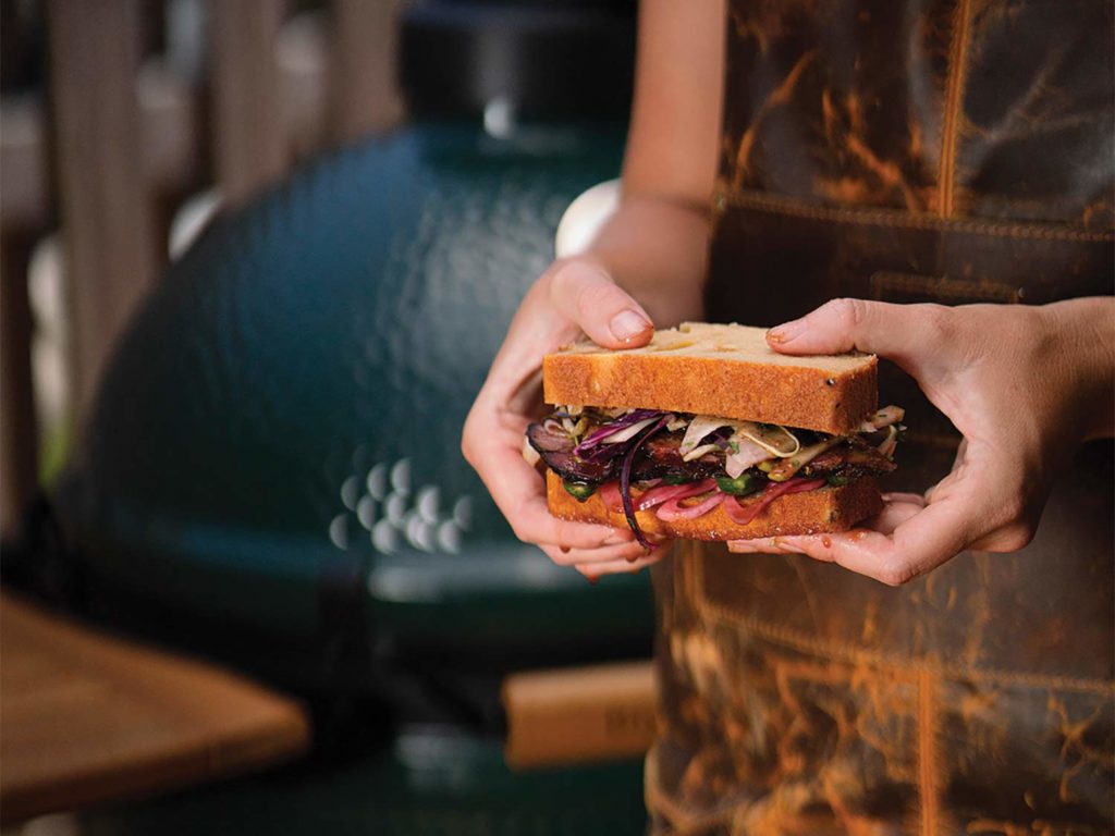 A hand holding a wild-game sandwich cooked on a Big Green Egg smoker.