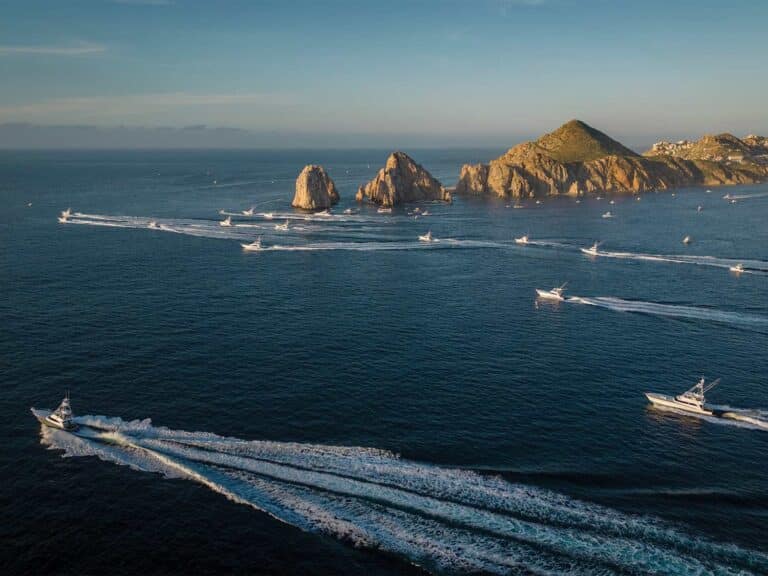 A fleet of sport-fishing boats cruise across the bay in Los Cabos. In the distance, the El Arco rocky outcrops sit aside their path.