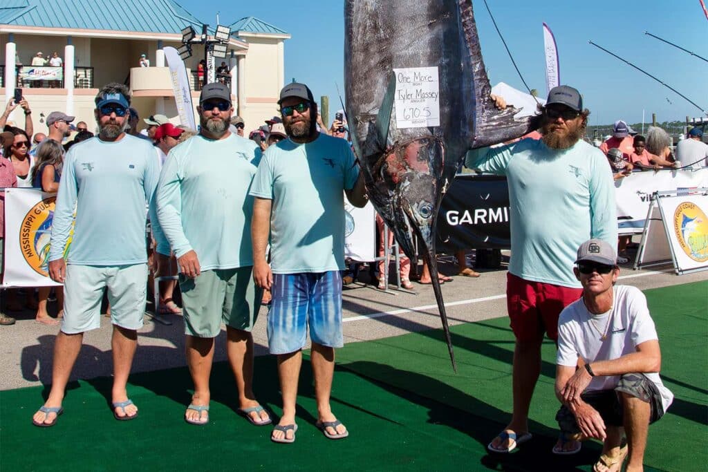 A team of four five men standing beside a strung up blue marlin at a tournament's weigh-in ceremony. They are surrounded by a large excited crown.