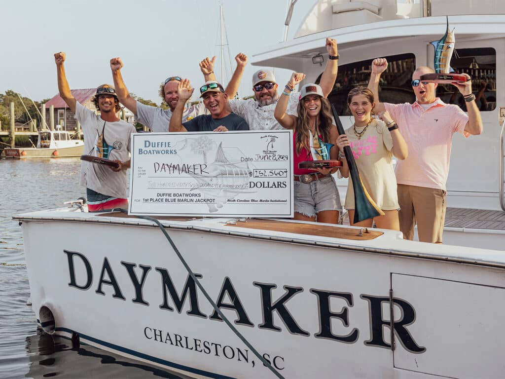 A sport-fishing team poses in celebration in the cockpit of their boat DayMaker. They hole their hands up with trophies and an oversized check.