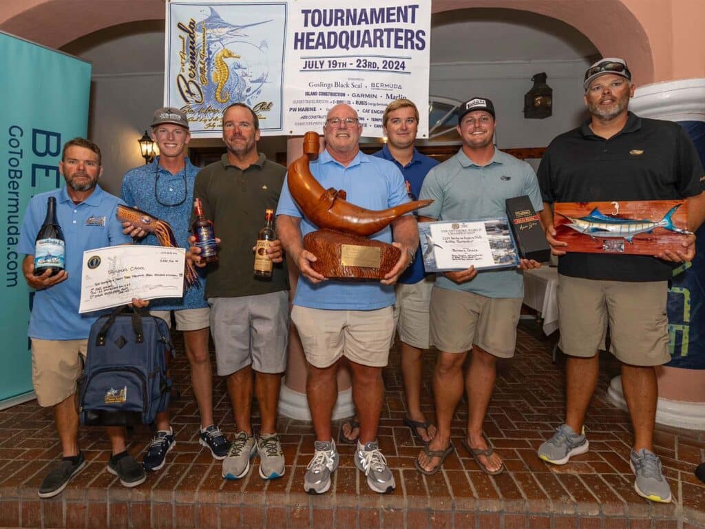 A team of sport-fishing anglers stand on stage at the Sea Horse Anglers Club Billfish Tournament. They hold up prizes, awards, over-sized checks. The center angler holds a wood-carved trophy in the shape of a seal.