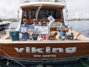 A sport-fishing team stands in the cockpit of the Viking 82 sport-fisher, celebrating their performance. In front of them are various prizes and trophies. The two center anglers hold up an oversized check.