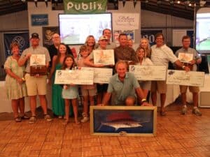 A large group of men and women, and their children celebrating victories at the 2024 Pirate's Cove Billfish Tournament. Several angelrs are holding plaques and awards. Several anglers are holding over-sized checks, and the center angler props up a piece of 3d sailfish artwork in a frame.