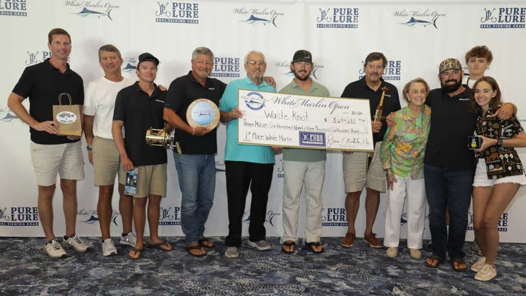 A lineup of a sport-fishing team at an awards ceremony. They are standing in front of a wall of logos, and holding an assortment of trophies, prizes, and an over-sized check.