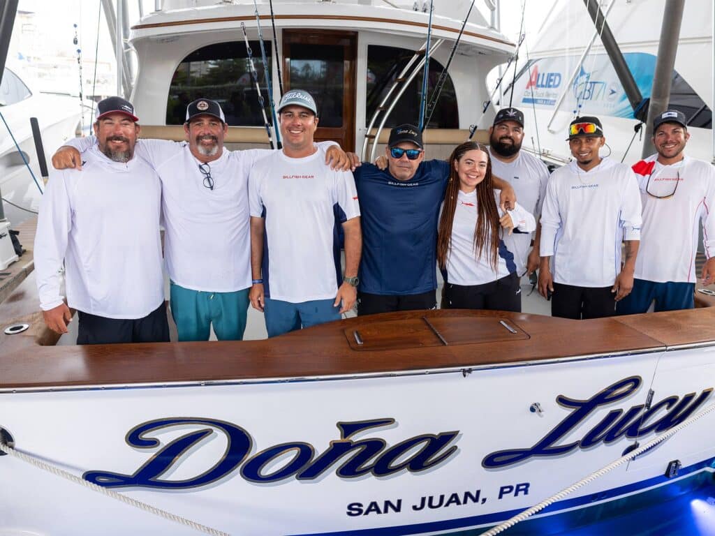 A crew of sport-fishing anglers standing in the cockpit of their sportfishing during the San Juan International Billfish Tournament. "Doña Lucy" is painted on the transom in script.