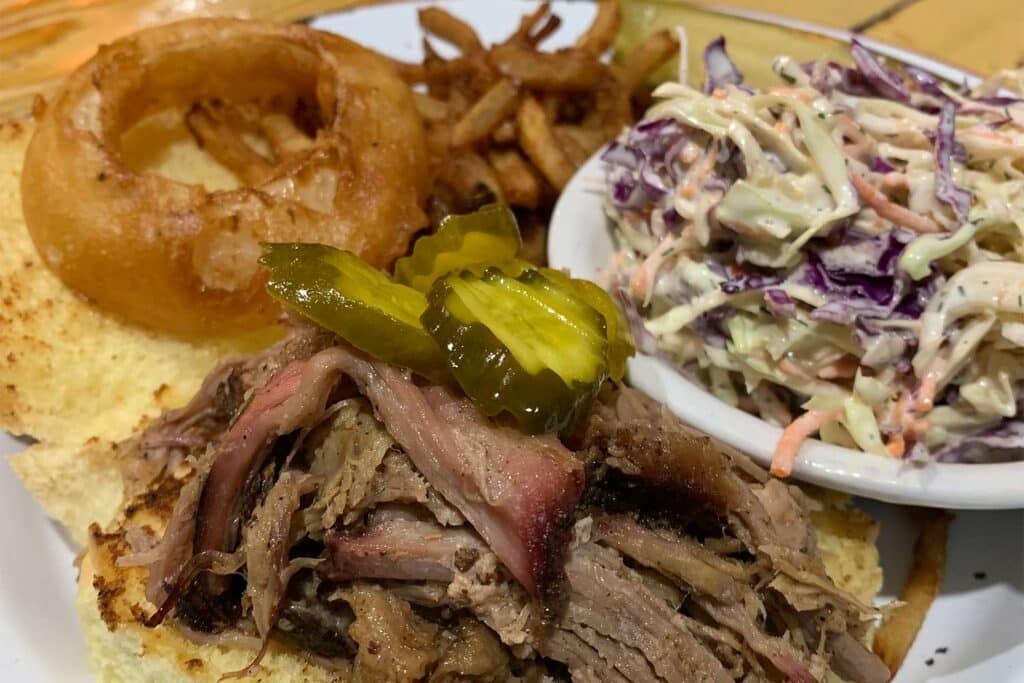 Close up detail of shreeded bbq next to a plate of onion rings and a bowl of slaw.