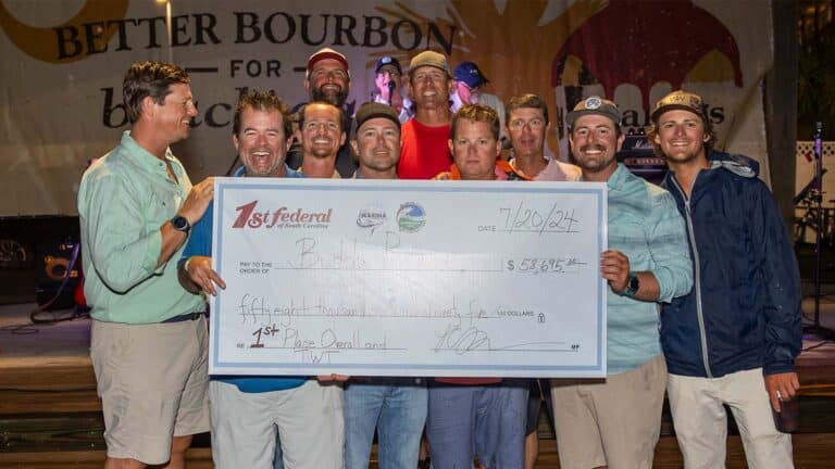 The team Buddy Rowe stand at an awards ceremony. They hold up an over-sized check in front of their group.