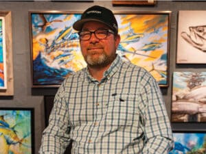 Dennis Friel sits and poses for the camera surrounded by his paintings on display in a gallery setting