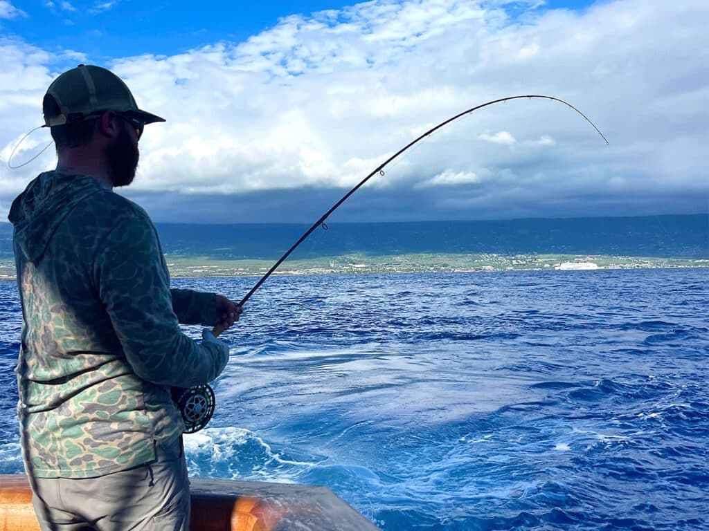 An angler stands in the cockpit of a sport-fishing boat, fishing. In the horizon you can see tropical coastline.