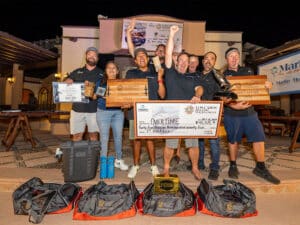 A sport-fishing team standing in celebration. They hold up awards and prizes and an over-sized check. Several other prizes are arranged on the ground around them.