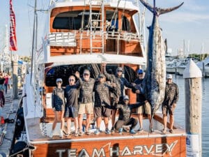 A sport-fishing team stands in the cockpit of the Team Harvey boat next to a large marlin being weighed at the docks.