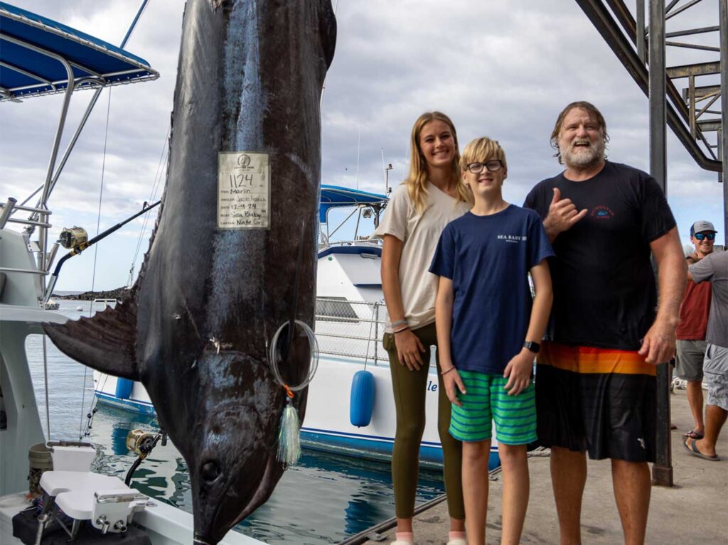 A fishing family stands next to a grander blue marlin being weighed dockside.